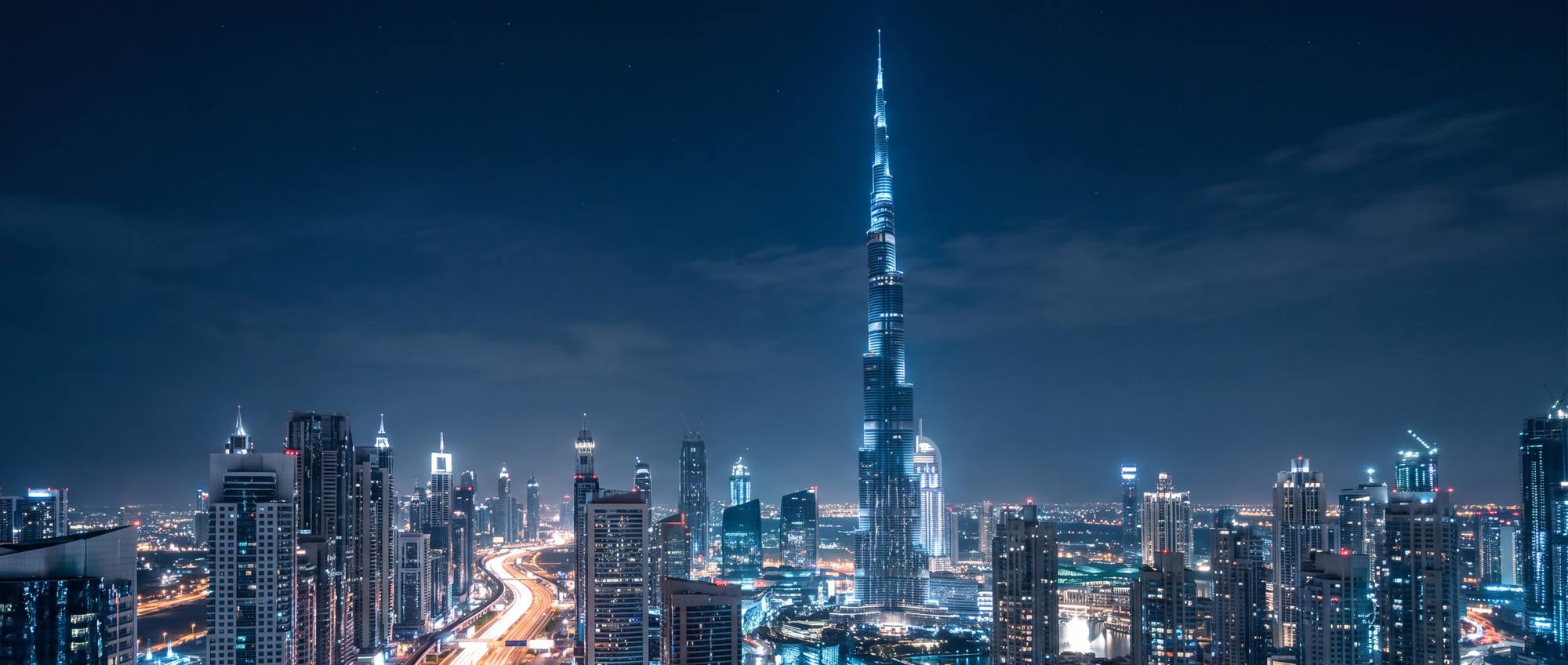The Dubai skyline at night, the Burj Khalifa lit above the towers of Sheikh Zayed Road with traffic light trails along the highway